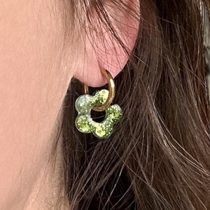 Pode incluir: Um par de brincos de argola dourados com um charme de flor verde e dourado com glitter. O charme da flor é um design simples com cinco pétalos.