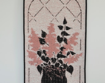 May include: A pink and black woven wall tapestry featuring a floral design with a vase and a window frame.