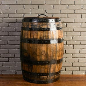 May include: A cylindrical wooden barrel featuring vertical staves with a natural brown finish. The barrel is reinforced by four horizontal black metal bands secured with rivets. A circular wooden lid with a small black metal handle sits on top.