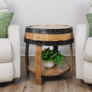 May include: A round side table constructed from a repurposed wooden barrel with a natural light brown finish. The table features two dark, weathered metal bands with visible rivets and a lower circular shelf holding a small white pot with greenery.