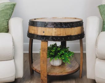 May include: A round side table constructed from a repurposed wooden barrel with a natural light brown finish. The table features two dark, weathered metal bands with visible rivets and a lower circular shelf holding a small white pot with greenery.
