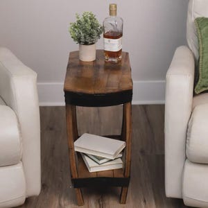Op de afbeelding: Een smalle bijzettafel gemaakt van gebogen houten tonduigen met zwarte metalen banden. De tafel heeft een plat bovenoppervlak met een groene plant in een pot en een glazen fles, en een onderste plank met twee op elkaar gestapelde witte boeken.