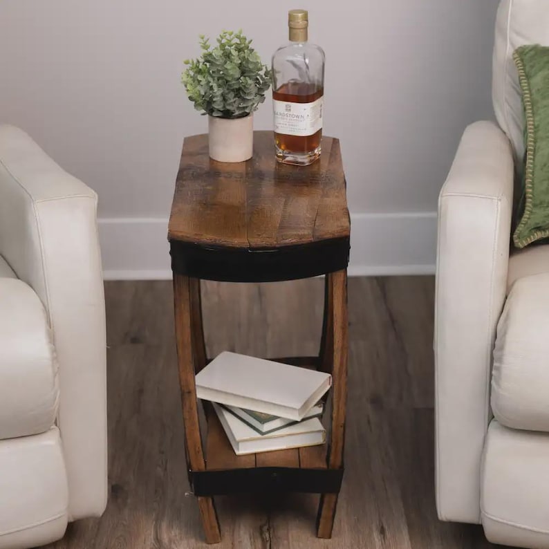 Op de afbeelding: Een smalle bijzettafel gemaakt van gebogen houten tonduigen met zwarte metalen banden. De tafel heeft een plat bovenoppervlak met een groene plant in een pot en een glazen fles, en een onderste plank met twee op elkaar gestapelde witte boeken.