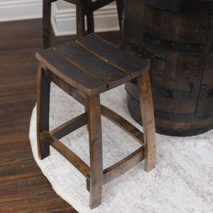 May include: A square stool constructed from dark brown wood staves with a charred black finish on the seat and legs. The stool features four curved legs connected by horizontal support braces, showcasing a rustic wood grain texture throughout.