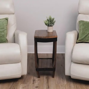 Op de afbeelding: Een hoge, smalle houten bijzettafel met een donkerbruine afwerking, voorzien van een rechthoekig blad en een onderste plank. De tafel heeft gebogen poten en staat onder een kleine, lichte pot met een groene kunstplant.