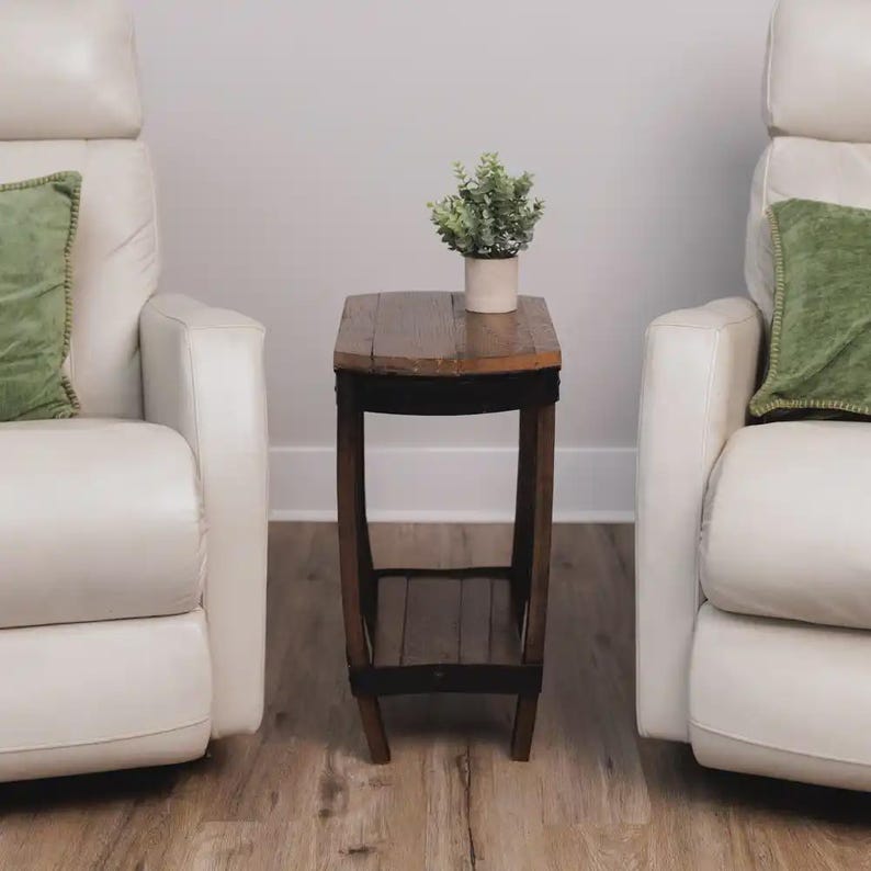 Op de afbeelding: Een hoge, smalle houten bijzettafel met een donkerbruine afwerking, voorzien van een rechthoekig blad en een onderste plank. De tafel heeft gebogen poten en staat onder een kleine, lichte pot met een groene kunstplant.