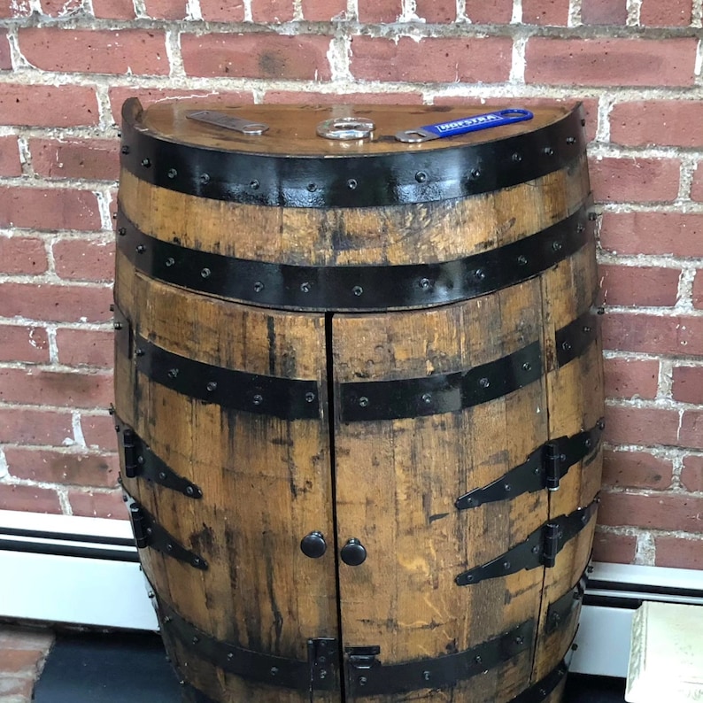 May include: A wooden barrel shaped cabinet with two doors. The cabinet is made of brown wood with black metal accents. The doors are closed and have black metal hinges. The cabinet is sitting in front of a brick wall. The top of the cabinet has a black metal trim and a blue metal bottle opener with the word 'Drinkfix' on it.