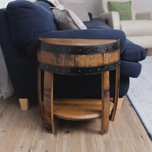 May include: A semi-circular side table constructed from a repurposed wooden barrel. The table features a natural wood grain finish, two horizontal black metal bands secured with rivets, and a lower shelf for storage, supported by curved legs.