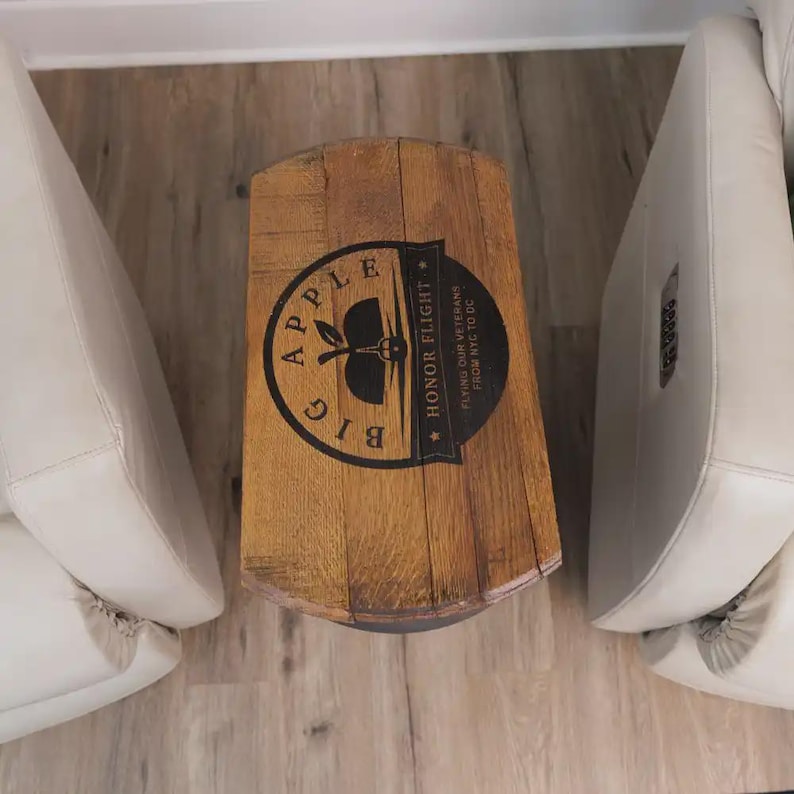 Op de afbeelding: Een cilindervormige houten bijzettafel met een natuurlijke bruine afwerking. Het blad is voorzien van een zwarte cirkelvormige afbeelding met een appelicoon en de tekst Big Apple Honor Flight Flying Our Veterans From NYC To DC.
