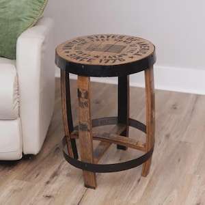 May include: A round wooden stool constructed from barrel staves with black metal bands. The top features a printed American flag graphic and the text UNITED STATES OF AMERICA LAND OF THE FREE HOME OF THE BRAVE SINCE 1776 in black ink.