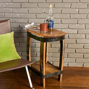 Op de afbeelding: Een bijzettafel gemaakt van een hergebruikt houten vat met zwarte metalen banden, voorzien van een gebogen tafelblad en een onderste plank. Op de tafel staan een glazen fles Weller bourbon en een glas met een bruine lederen hoes.