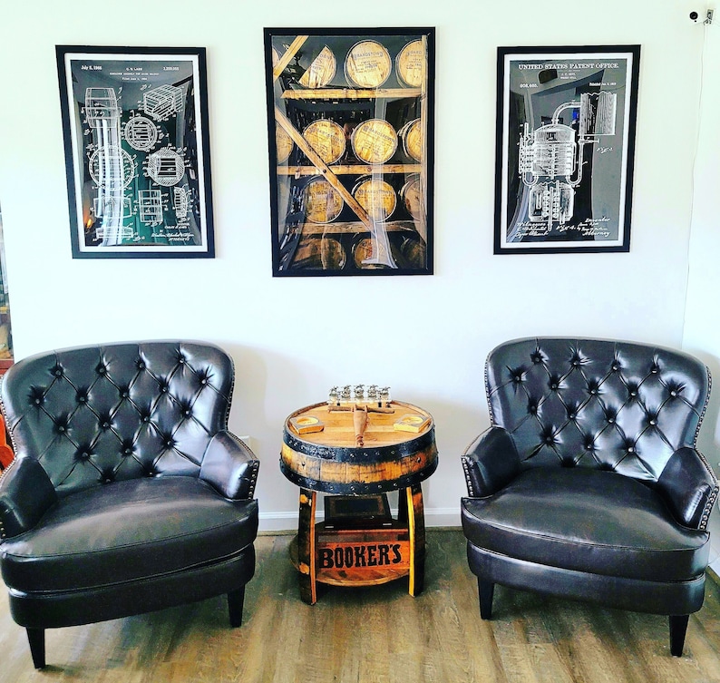 May include: Two black leather armchairs with tufted backs and a wooden barrel table with a sign that reads "Booker's" in front of three framed black and white prints. The prints are of vintage diagrams of a distillery.