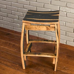 May include: A stool constructed from curved wooden barrel staves featuring a natural light brown wood grain finish and black painted accents on the seat and legs. The structure includes four curved legs connected by horizontal support braces.