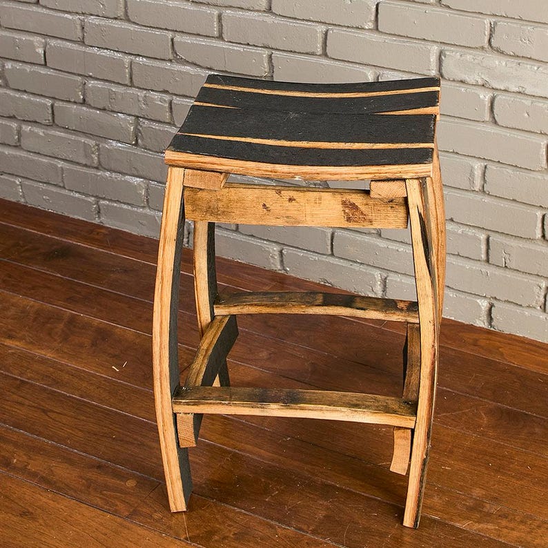 May include: A stool constructed from curved wooden barrel staves featuring a natural light brown wood grain finish and black painted accents on the seat and legs. The structure includes four curved legs connected by horizontal support braces.