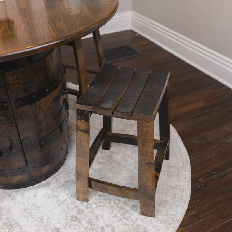 May include: A square wooden stool with a dark brown finish, featuring four sturdy legs and a slatted seat. The stool is constructed from reclaimed wood with visible grain patterns and metal hardware accents, matching the adjacent barrel-style table.