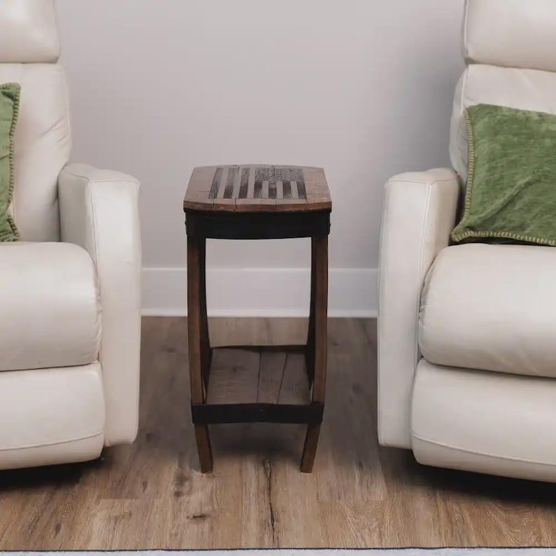 Op de afbeelding: Een kleine houten bijzettafel met een rechthoekig blad voorzien van een donker, gegraveerd Amerikaans vlagontwerp. De tafel heeft vier poten verbonden door een onderste plank en is gemaakt van donker, verweerd hout met zwarte metalen accenten.