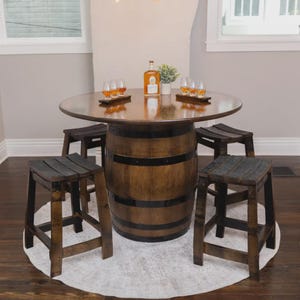 May include: A round wooden barrel table with black metal hoops and a smooth top, surrounded by four matching wooden stools. On the table sits a glass bottle of amber liquid, two small wooden serving trays with glasses, and a small potted plant.