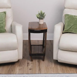 Op de afbeelding: Een hoge, smalle houten bijzettafel met een donkerbruine afwerking, voorzien van een rechthoekig blad en een onderste plank. Een kleine groene plant in een pot staat op het tafelblad, geplaatst tussen twee cr&egrave;mekleurige fauteuils.