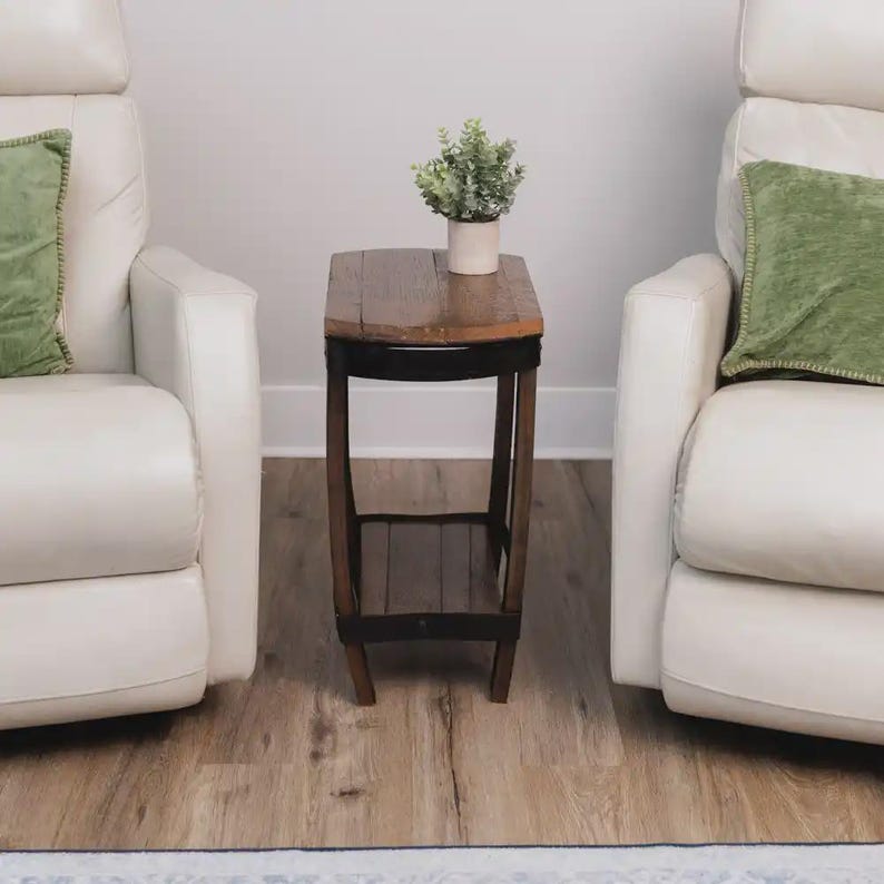 Op de afbeelding: Een hoge, smalle houten bijzettafel met een donkerbruine afwerking, voorzien van een rechthoekig blad en een onderste plank. Een kleine groene plant in een pot staat op het tafelblad, geplaatst tussen twee cr&egrave;mekleurige fauteuils.