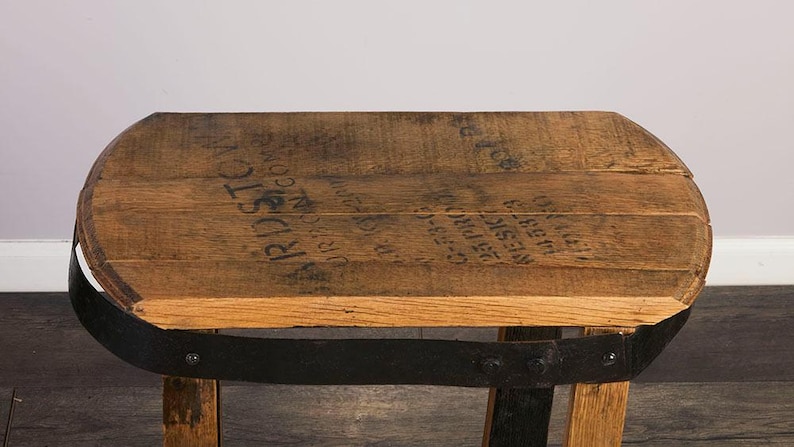 Op de afbeelding: Een kleine kruk gemaakt van een gerecyclede houten ton, met een natuurlijke eikennerf afwerking en een donkere metalen band rondom de zitting. Het bovenoppervlak toont zwarte gestencilde tekst met BARDSTOWN en andere markeringen.