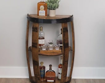 May include: A semi-circular wooden barrel shelf featuring a dark brown finish with black metal bands. The unit has two interior shelves holding various glass liquor bottles and three clear glasses. A small potted green plant sits on the top surface.