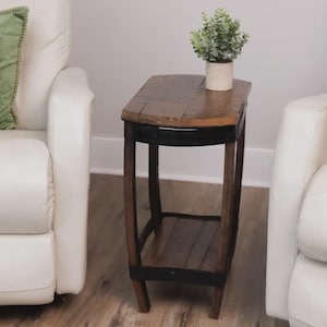 Op de afbeelding: Een hoge, smalle bijzettafel gemaakt van donkerbruine houten planken met een gebogen, tonvormig frame versterkt door zwarte metalen banden. De tafel heeft een vlak bovenoppervlak en een onderste plank met een kleine groene plant.