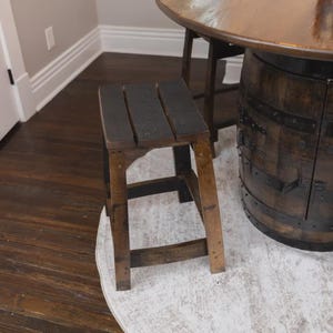 May include: A square wooden stool featuring a seat made of three dark, charred planks. The four legs are connected by horizontal support bars, showcasing a natural wood grain finish with dark, weathered accents throughout the structure.