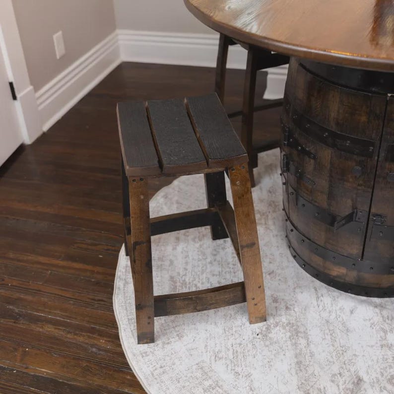 May include: A square wooden stool featuring a seat made of three dark, charred planks. The four legs are connected by horizontal support bars, showcasing a natural wood grain finish with dark, weathered accents throughout the structure.