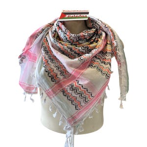 Könnte beinhalten: Gemustertes Tuch in Weiß, Rosa und Schwarz mit Quasten. Das Tuch hat ein geometrisches Design mit rosa und schwarzen Zickzackmustern. Das Etikett lautet "Product Of Palestine" und "Baladna".