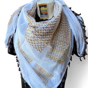 Könnte beinhalten: Weißer und goldfarbener Schal mit braunen Quasten. Der Schal hat ein geometrisches Muster und ist über einer schwarzen Form drapiert. Der Schal hat ein Etikett mit der Aufschrift "Visit Palestine".
