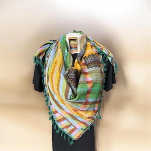 Könnte beinhalten: Ein farbenfrohes Keffiyeh-Tuch mit einer Mischung aus Mustern und Farben, darunter Grün, Gelb, Braun und Blau. Der Schal hat einen Fransenrand und ist über einem schwarzen Kleidungsstück drapiert. Das Etikett lautet "Palestine".