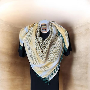 Könnte beinhalten: Ein gelb-weißes Keffiyeh-Tuch mit grünem Zickzackmuster und grüner Quastenborte. Der Schal ist über einer schwarzen Form drapiert. Der Schal hat ein Etikett mit der Aufschrift "Palestine".