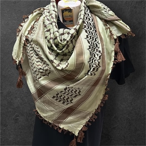 Può includere: Sciarpa keffiyeh beige e marrone con motivo geometrico e nappe marroni. La sciarpa è drappeggiata su una forma nera, mostrando il suo design e la sua consistenza. La sciarpa ha un design tradizionale mediorientale.