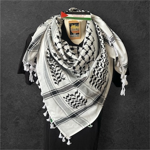 May include: A white and black keffiyeh scarf with a geometric pattern and tassel edges. The scarf is draped over a black shirt, with a tag that reads "Products Of Palestine".