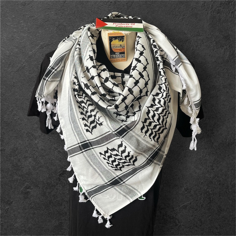 May include: A white and black keffiyeh scarf with a geometric pattern and tassel edges. The scarf is draped over a black shirt, with a tag that reads "Products Of Palestine".