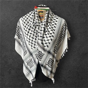 May include: A white and black keffiyeh scarf with a geometric pattern and fringed edges. The scarf has a gray border and a tag with text and a flag. The scarf is draped over a black form.