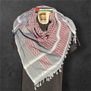 Könnte beinhalten: Ein grau-rotes Keffiyeh-Tuch mit geometrischem Muster und Quastenbesatz. Das Tuch ist über einem schwarzen Kleidungsstück drapiert, mit einem Etikett mit der palästinensischen Flagge und den Worten "Palestine" und "Products of Palestine".