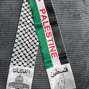 May include: A white scarf with black trim and fringe. The scarf features the Palestinian flag with the words "Palestine" and "فلسطين" printed on it. The scarf also features a black and white image of the Dome of the Rock and the text "القدس لنا" printed on it.