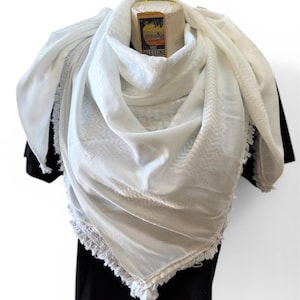 Könnte beinhalten: Ein weißes Keffiyeh-Tuch mit geometrischem Muster und Fransen. Der Schal ist über einem schwarzen Kleidungsstück drapiert. Ein kleines Etikett mit dem Text "Visit Palestine" ist sichtbar.