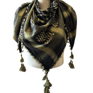May include: A black and olive green keffiyeh scarf with a checkered pattern and decorative tassels. The scarf is draped over a mannequin and features the text "Baladna" on a tag.