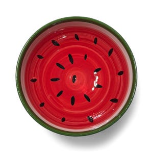 Pode incluir: Uma tigela redonda de cerâmica com um design de melancia. O interior é vermelho brilhante com formas de sementes pretas e a borda é verde. A tigela está sobre um fundo branco.