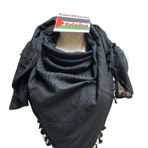 Könnte beinhalten: Ein schwarzes Keffiyeh-Tuch mit strukturiertem Muster und Quastenbesatz. Das Tuch ist über einem Schaufensterpuppenkopf drapiert. Ein Etikett oben zeigt "Product Of Palestine" und "Baladna" mit einer palästinensischen Flagge.