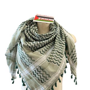 Könnte beinhalten: Ein grün-weißer Keffiyeh-Schal mit geometrischem Muster und Fransenquasten. Der Schal ist über einem Schaufensterkopf drapiert. Das Etikett lautet "Product Of Palestine" und "Baladna".