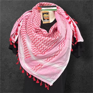 Könnte beinhalten: Ein rosa-weißer Keffiyeh-Schal mit roten Zickzackmustern und Quasten. Der Schal ist über einem schwarzen Kleidungsstück drapiert, mit einem Etikett mit der Aufschrift "Products of Palestine".