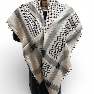 May include: A beige and black keffiyeh scarf with a geometric pattern and fringed edges. The scarf is draped over a black form, showcasing its design. The scarf has a tag that says "Palestine".
