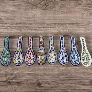 May include: A collection of ceramic spoon rests with colorful floral and architectural designs. The spoon rests feature a variety of patterns, including flowers, buildings, and olive branches, with a blue, red, yellow, and green color palette.