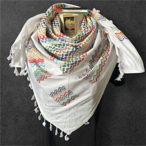 Könnte beinhalten: Weißes Keffiyeh-Tuch mit bunten geometrischen Mustern und Fransenbesatz. Das Tuch hat ein traditionelles Design mit roten, grünen, blauen und gelben Akzenten. Ein kleines Etikett mit dem Text "Visit Palestine" ist angebracht.