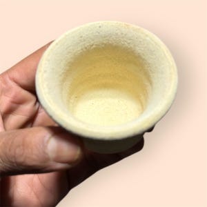 May include: A small, off-white ceramic cup with a rounded rim and a pedestal base. The interior of the cup has a light yellow hue. The cup is held in a hand, with the opening facing upwards.