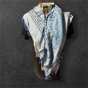 Könnte beinhalten: Ein hellblau-weißer Keffiyeh-Schal mit blauen Quasten. Der Schal hat ein geometrisches Muster in Blau und Schwarz. Der Schal ist über einem schwarzen T-Shirt drapiert.