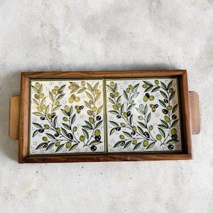 May include: A rectangular wooden serving tray with two ceramic tiles featuring olive branch designs. The tiles have a white background with green leaves and black and green olives. The tray has wooden handles.